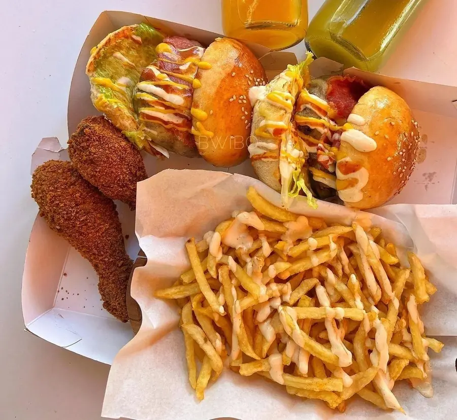 Two burgers in paper boxes, fried chicken drumsticks, fries with sauce and two bottles of Bwibo Juice, from above