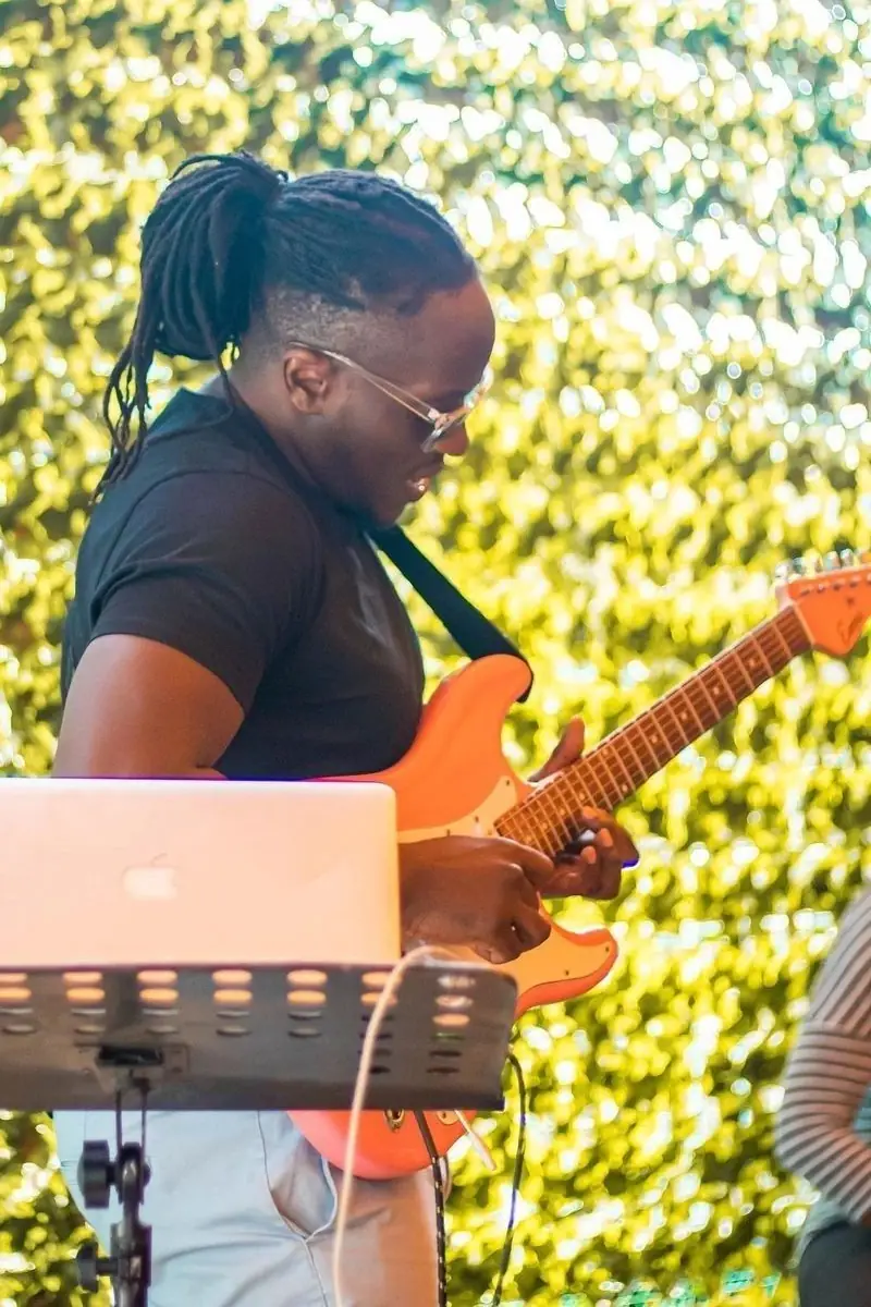 A guitarist playing in daylight in front of Bwibo’s green hedge wall