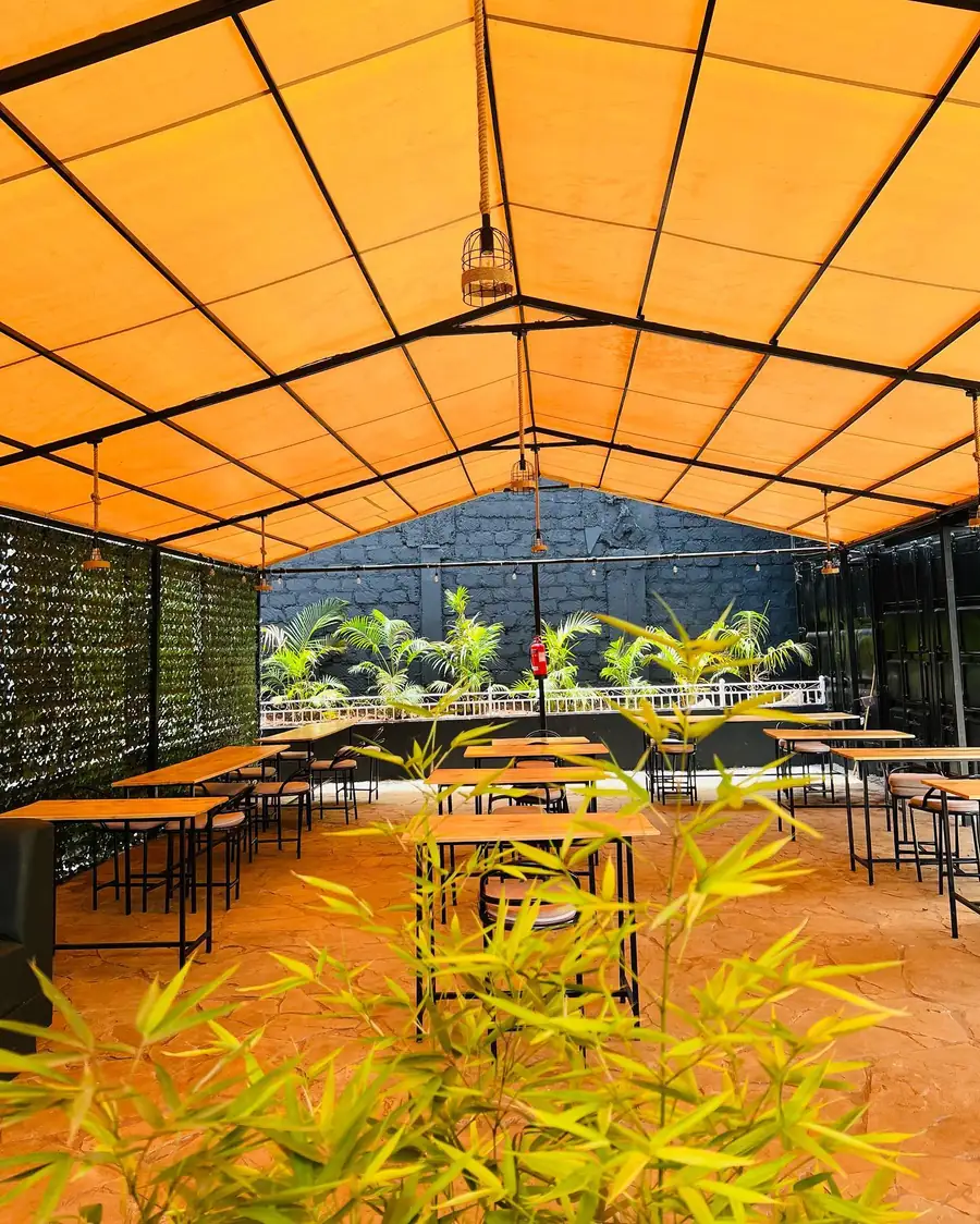 The dining area under Bwibo’s orange canopy: light-wood high tables, caged pendant lights, palms against a black stone wall